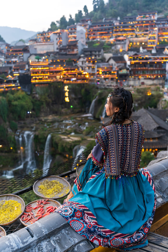 Una mujer con vestimenta tradicional contempla de espaldas un pintoresco pueblo antiguo iluminado junto a una cascada