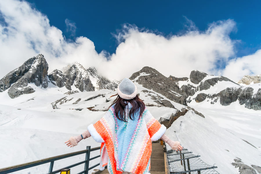 Una viajera con un colorido poncho y gorro de lana contempla de espaldas los picos nevados de la Montaña Nevada del Dragón de Jade