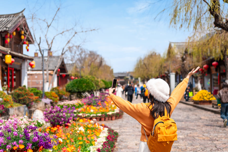 Viajera con mochila amarilla posa con los brazos abiertos en una calle empedrada del Casco Antiguo de Lijiang, rodeada de flores.